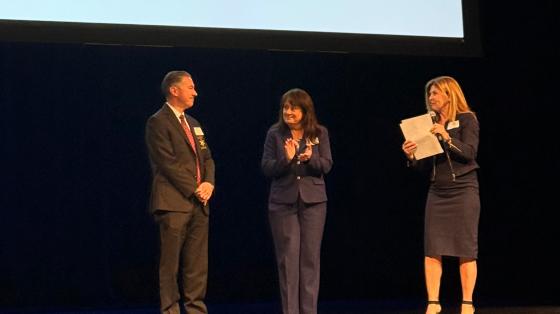 Three people stand on a stage in front of a projection screen during a presentation
