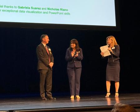 Three people stand on a stage in front of a projection screen during a presentation